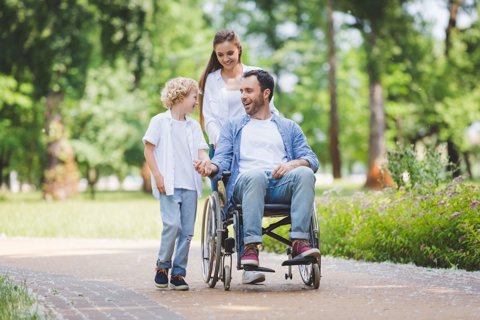 A young family enjoying the outdoors. The father is in a wheelchair, being pushed by his wife while he holds his son's hand
